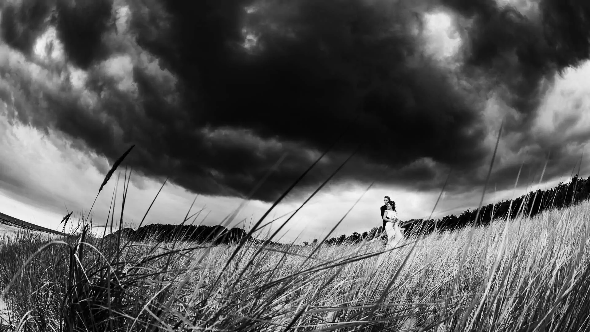Dramatisches Hochzeitsfotoin Schwarz Weiß. Brautpaar in den Dünen am Meer. Weitwinkel, dunkle Wolken